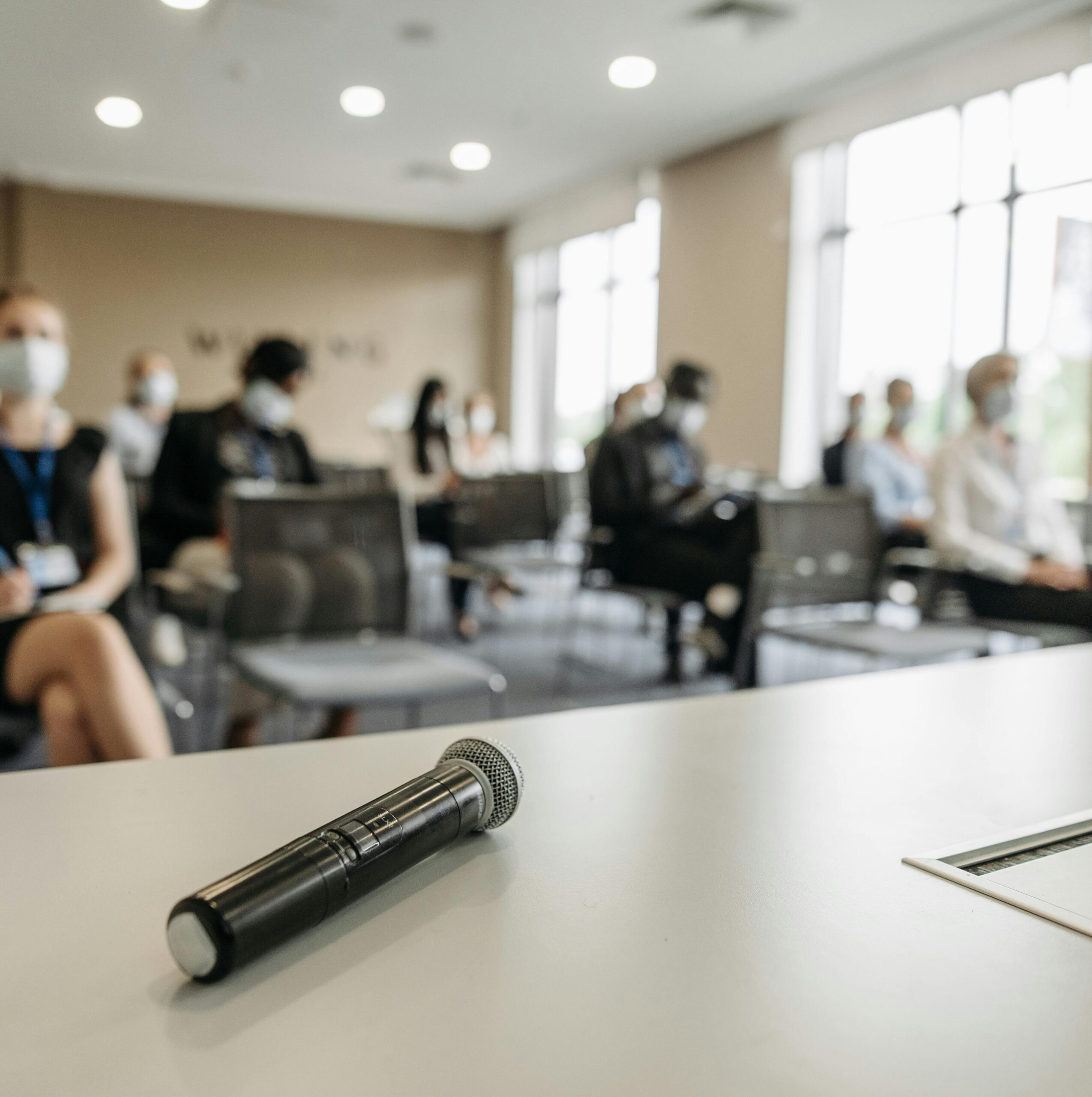 Im Vordergrund befindet sich ein Mikrofon auf einem Tisch. Im Hintergrund sieht man einen mit Menschen gefüllten Konferenzraum.