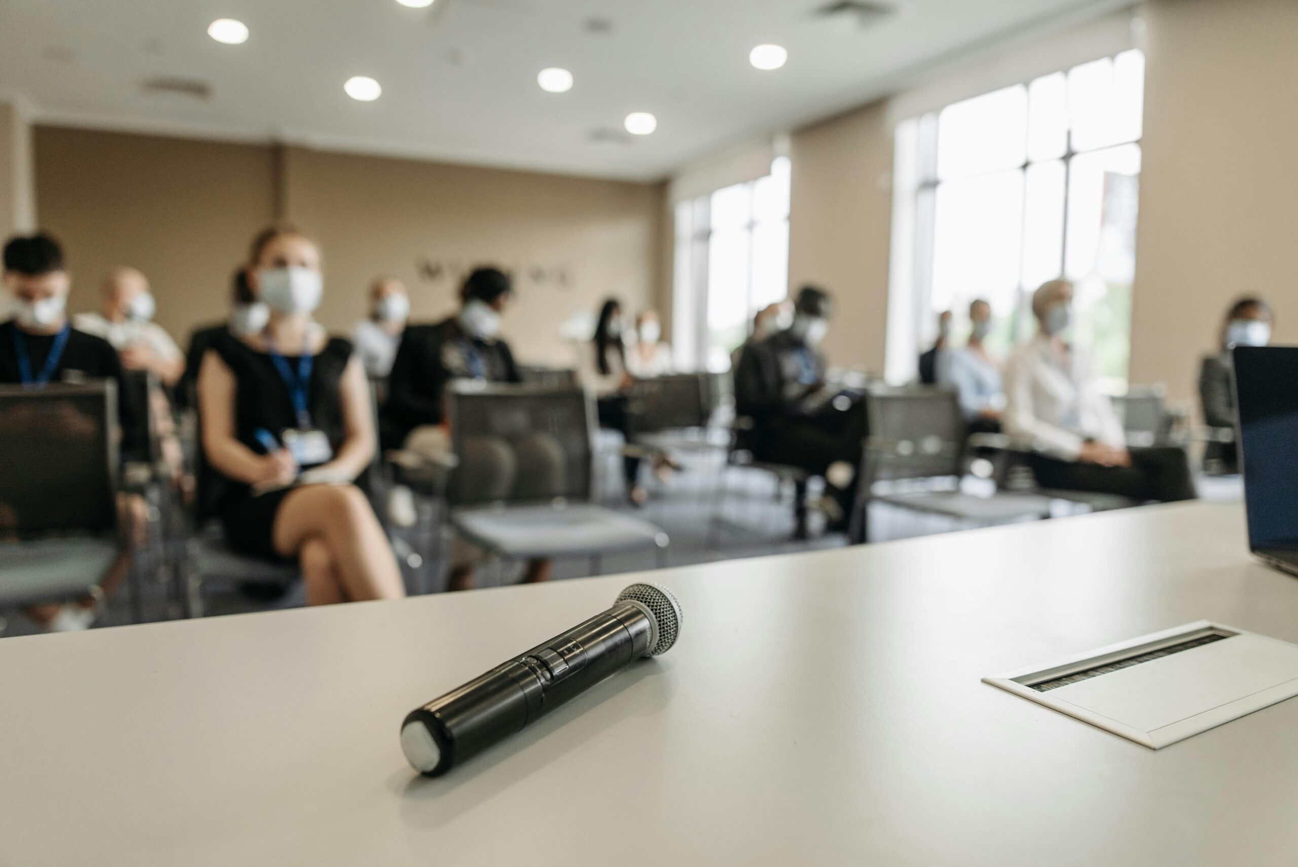 Im Vordergrund befindet sich ein Mikrofon auf einem Tisch. Im Hintergrund sieht man einen mit Menschen gefüllten Konferenzraum.