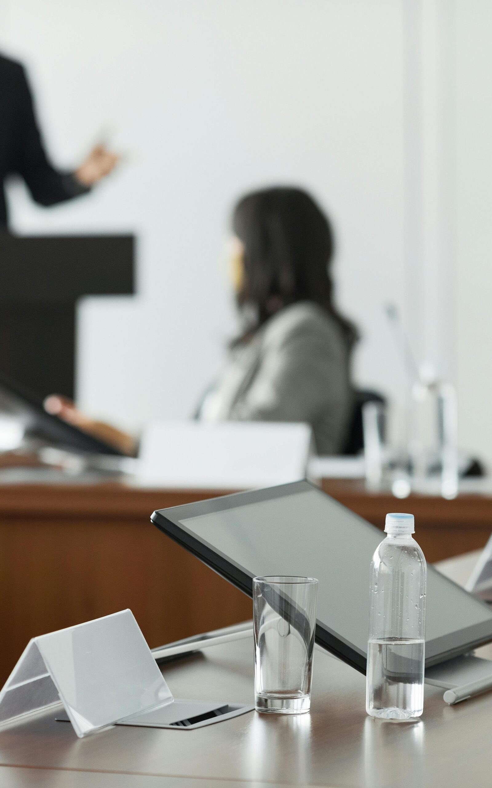 Es ist der Ausschnitt eines Konferenzsaals zu sehen. Im Vordergund ist ein Tablet, sowie eine Wasserflasche mit einem Trinkglas. Im Hintergrund befindet sich eine sitzende Frau, die sich mit einer weiteren Person außerhalb des Bildes unterhält.