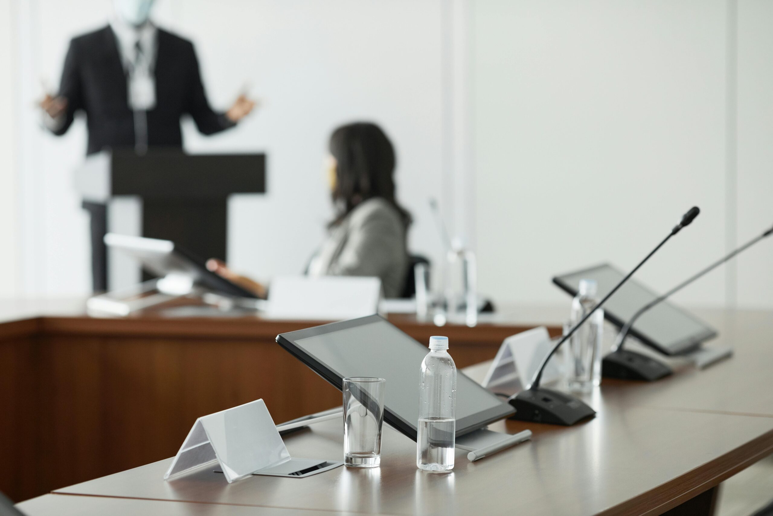 Es ist der Ausschnitt eines Konferenzsaals zu sehen. Im Vordergund ist ein Tablet, sowie eine Wasserflasche mit einem Trinkglas. Im Hintergrund befindet sich eine sitzende Frau, die sich mit einer weiteren Person außerhalb des Bildes unterhält.