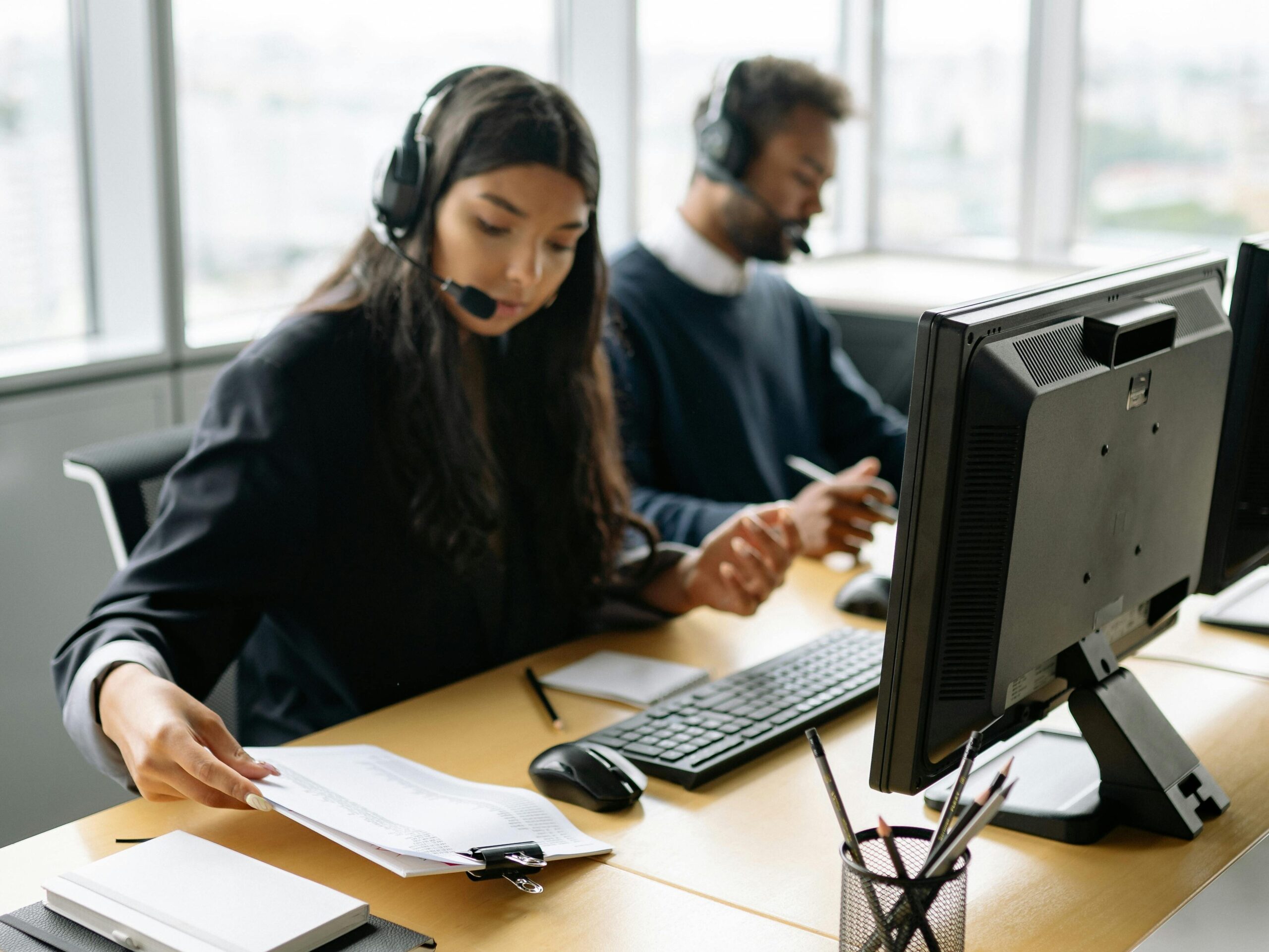 Eine Frau sitzt im Büro vor einem Comupter, berät jemanden über ein Headset und überprüft dabei ein Dokument.
